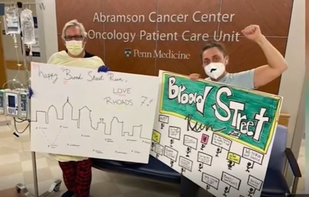 A male and female patient stand at the Abramson Cancer Center Oncology Patient Care Unit holding up hand-made posters that say “Broad Street Run” and “Happy Broad Street Run, LOVE RHOADS 7!”
